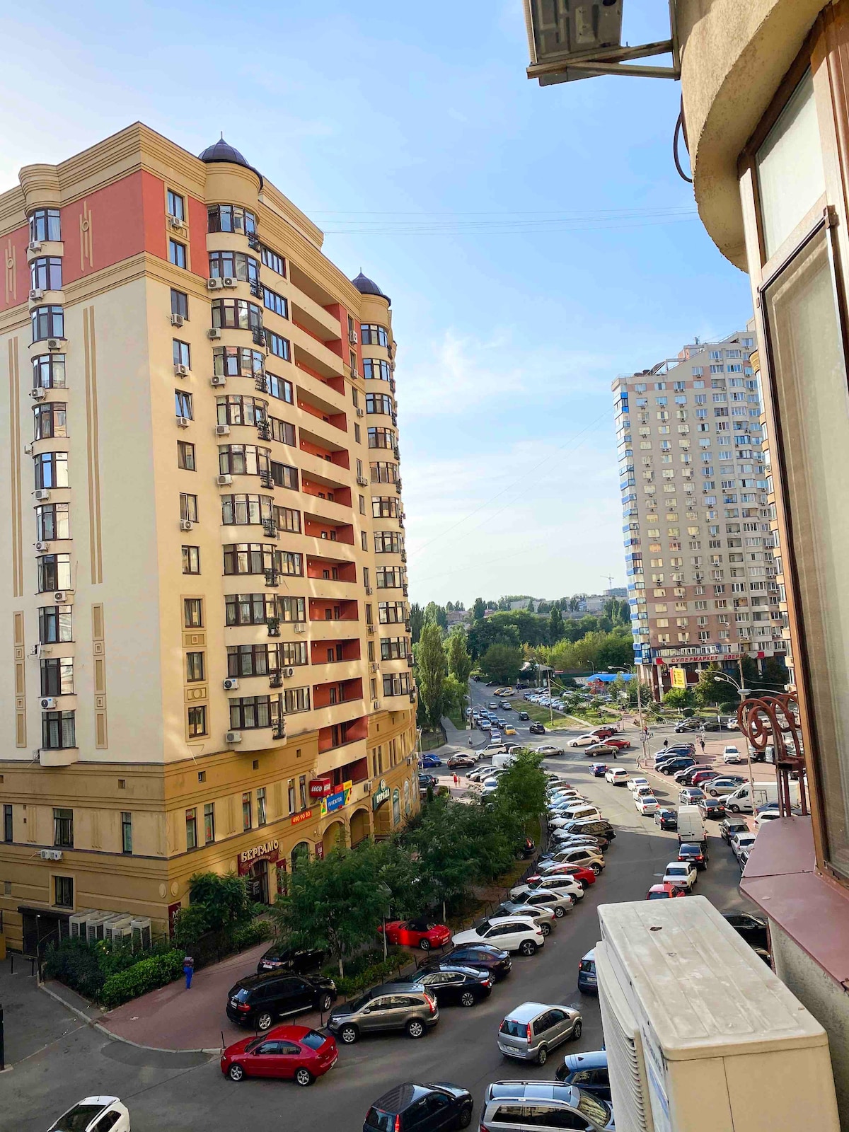 A view from the apartment reveals a busy city street lined with parked cars. The surrounding buildings exhibit diverse architectural styles, showcasing a blend of classic and modern design. Lush greenery is visible, providing a contrast to the urban environment.
