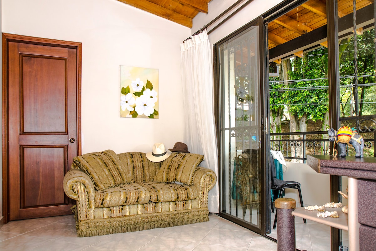 A living area features a plush, fringed sofa adorned with decorative pillows. Natural light filters through large glass doors that open to a balcony, framed by sheer curtains. A painting of white flowers decorates the wall, adding a touch of color to the room.