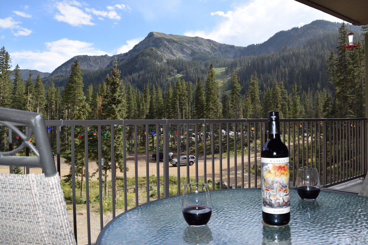 A scenic covered deck presents a table with a bottle of wine and two glasses, set against a backdrop of lush mountains and pine trees. The expansive view showcases ski slopes and rugged terrain, providing a tranquil outdoor space for relaxation.