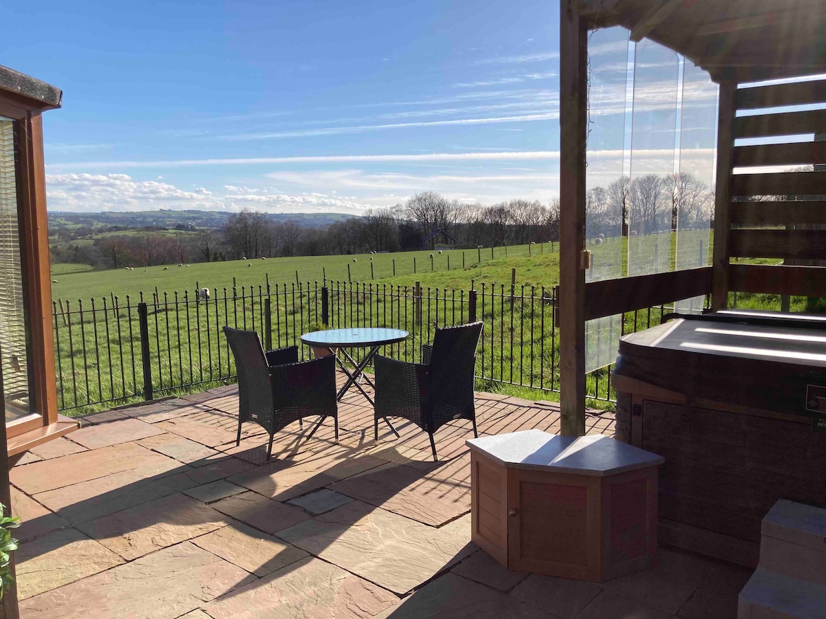 A sunlit patio is visible, featuring a round glass table surrounded by two wicker chairs. In the foreground, a wooden hot tub is positioned beside the outdoor seating. The landscape beyond is marked by rolling fields under a clear blue sky.