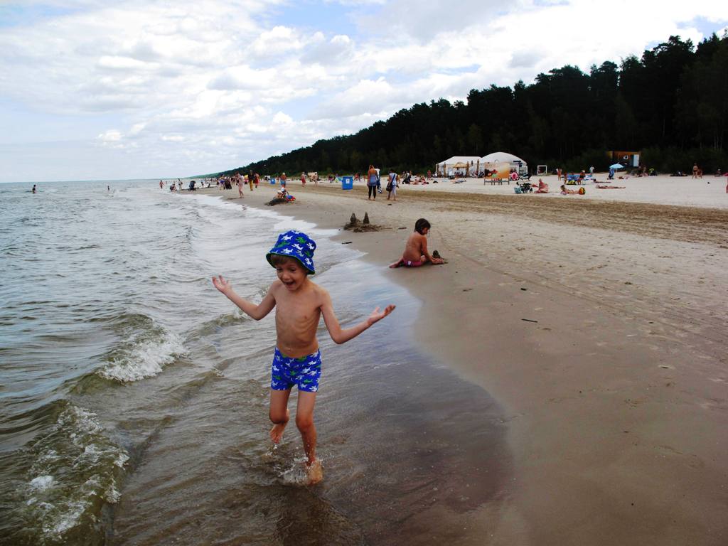 A sandy beach is lined with people enjoying the sun and water. Gentle waves lap at the shore, where a child in a blue hat and swim shorts plays. In the background, sunbathers and beach tents are visible against a backdrop of pine trees.