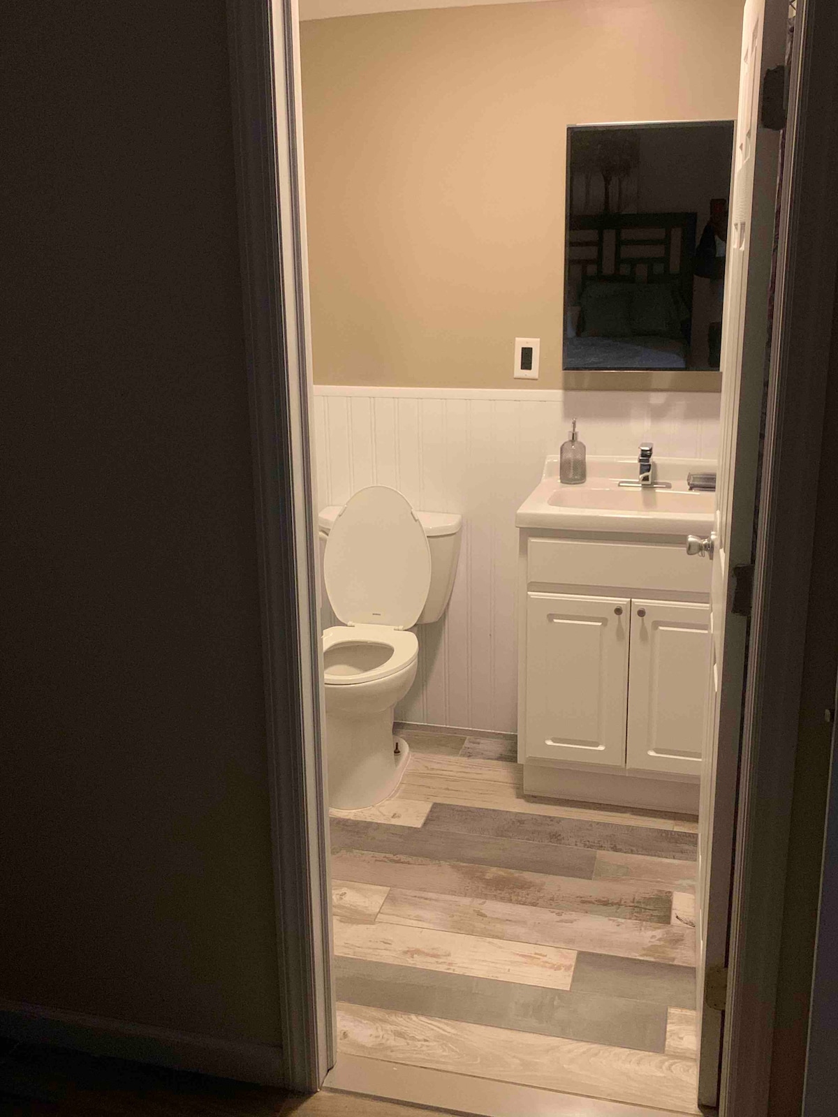 A small bathroom is visible, featuring a clean white vanity with a single sink and a large mirror above it. Light beige walls contrast with the light-colored wooden flooring. A toilet is positioned next to the vanity, creating a simple and functional layout.