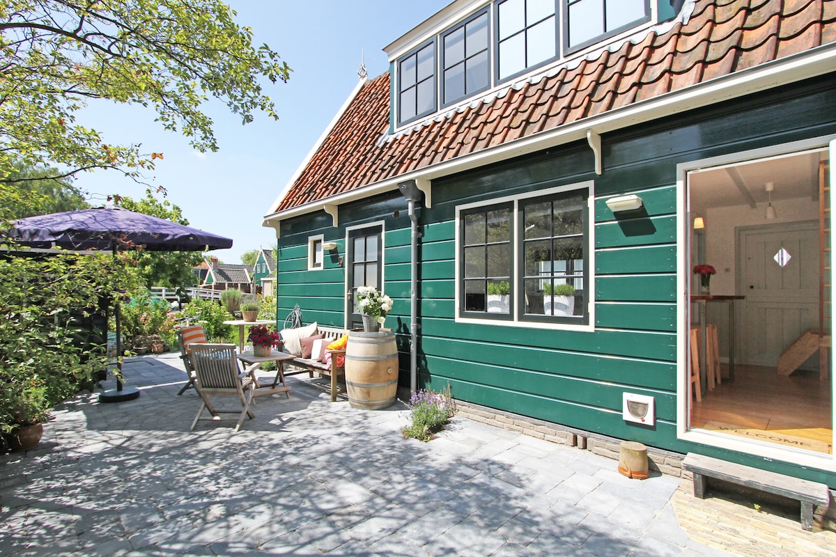 The exterior of the green wooden house is showcased, featuring a spacious terrace with seating arrangements and an umbrella. Potted plants add greenery to the space, while large windows offer natural light to the interior. A tranquil atmosphere is highlighted by the surrounding village scenery.