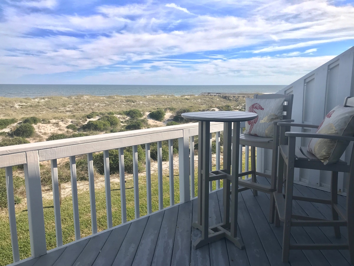A balcony features a bar-height table and two chairs, positioned to offer a clear ocean view. The deck is framed by white railings and overlooks sandy dunes, with the sea visible in the distance under a partly cloudy sky.