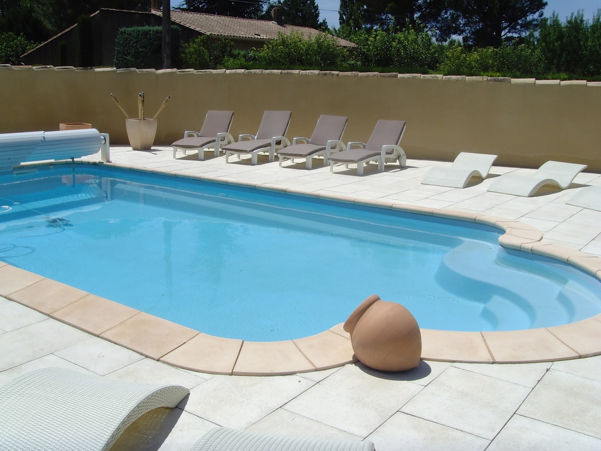 A shared outdoor pool area is visible, featuring light-colored stone tiles surrounding the clear blue water. Several lounge chairs are positioned for relaxation, with a few sun loungers placed for comfort. Greenery is present in the background, providing a natural setting.