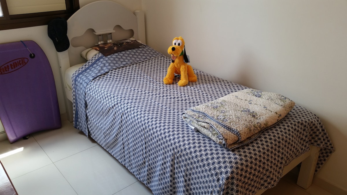 A single bed is positioned against a wall, covered with a patterned blue and white spread. A yellow stuffed toy sits atop the bed, alongside a neatly folded blanket. A lavender suitcase is placed in the corner, and natural light is visible through a nearby window.