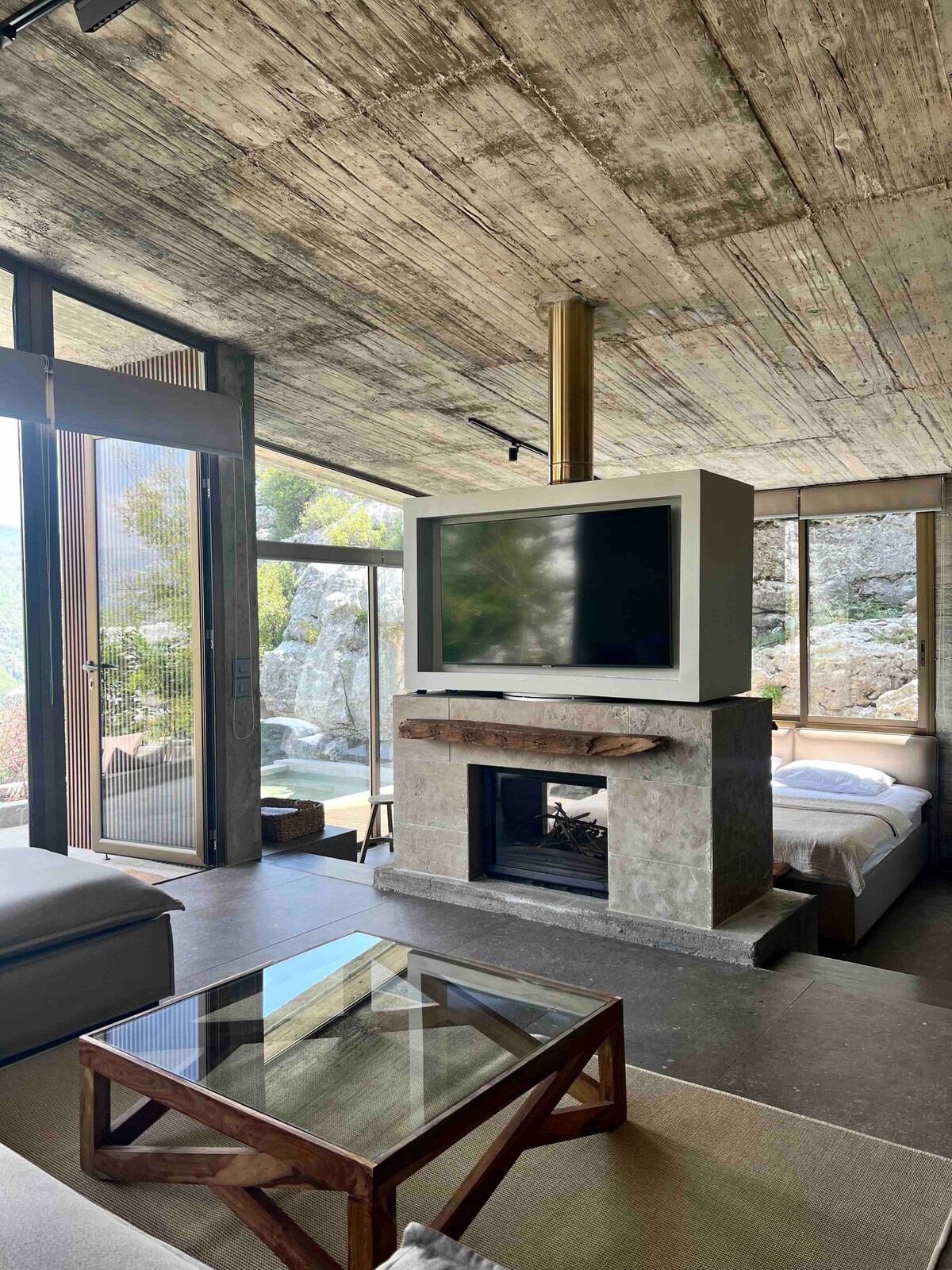 A cozy living area features a wood-burning fireplace positioned beneath a modern television. Large glass doors provide expansive views of the surrounding nature. A spacious sofa and glass coffee table complement the relaxed aesthetic, while natural light brightens the room through the windows.