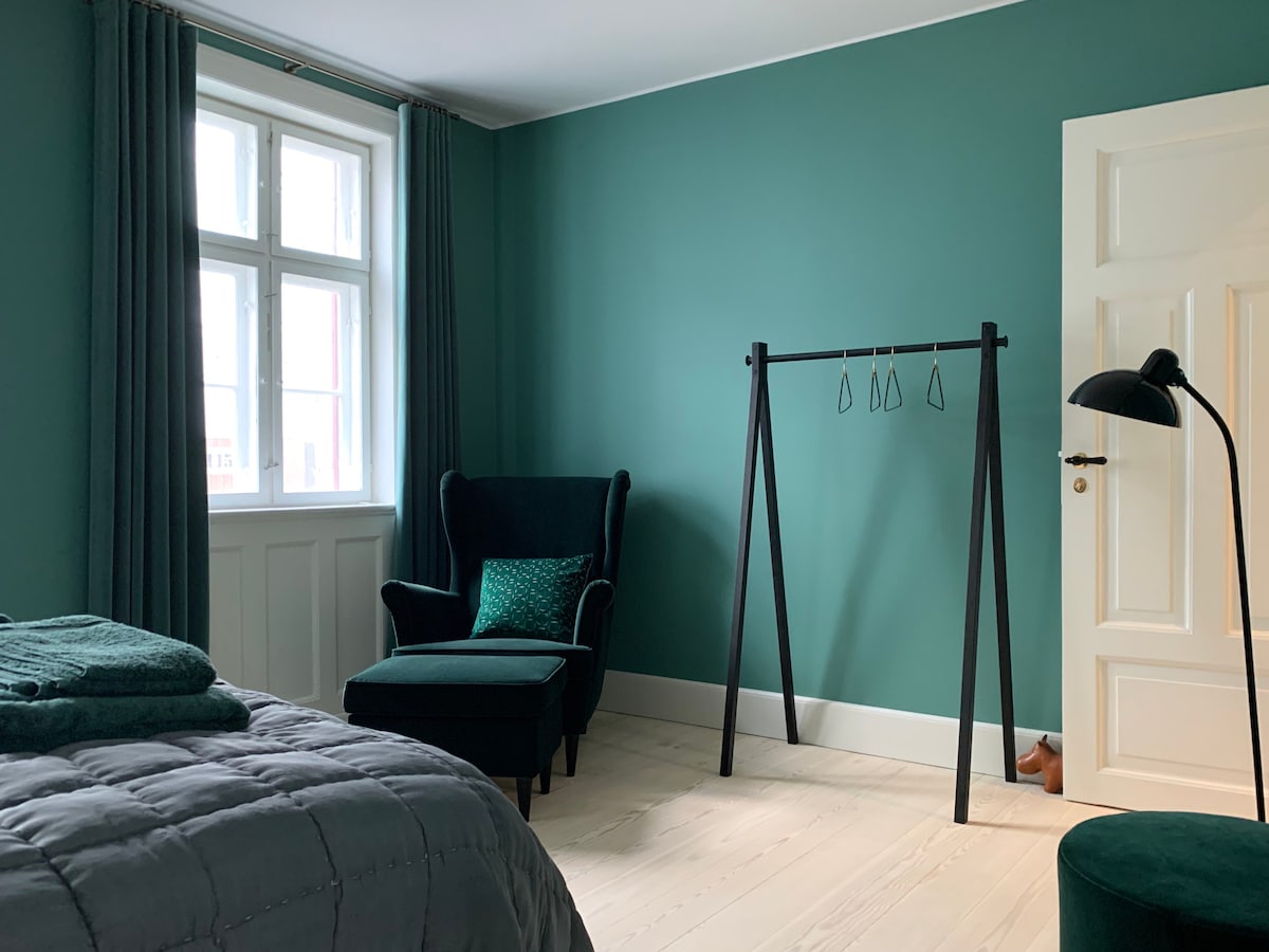 A spacious bedroom features a calming green wall and wooden floors. A cozy armchair sits beside a minimalist clothing rack, while a sleek floor lamp provides soft illumination. Natural light enters through a window adorned with matching curtains.