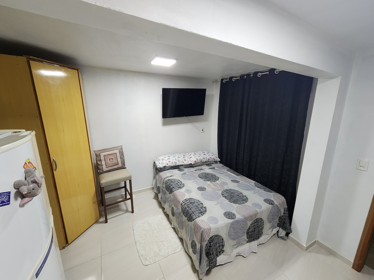 A cozy room is presented, featuring a double bed covered with a patterned blanket. A television is mounted on the wall across from the bed. A wooden chair and a storage cabinet are positioned nearby, while dark curtains provide privacy.