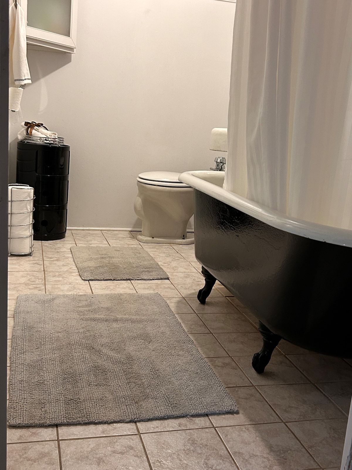 A modern bathroom featuring a classic clawfoot tub with black exterior. Light gray bath mats are positioned on a tiled floor. An illuminated white wall contrasts with the dark tub, while a toilet and toiletries in a storage unit are visible in a neat arrangement.