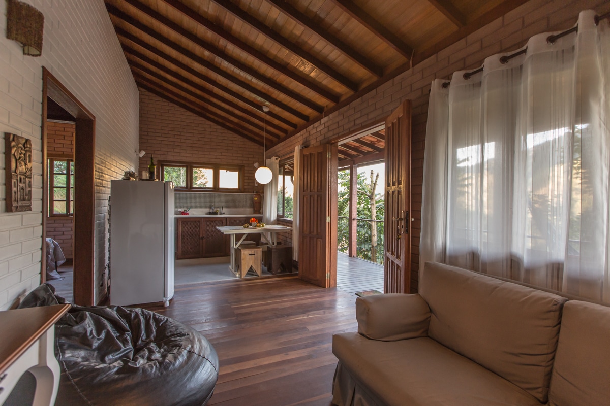 The open-plan interior features a cozy living area with a light-colored sofa and a bean bag. A dining table is positioned near the kitchen, which includes a white fridge. Large wooden doors lead outside, allowing natural light to fill the space.