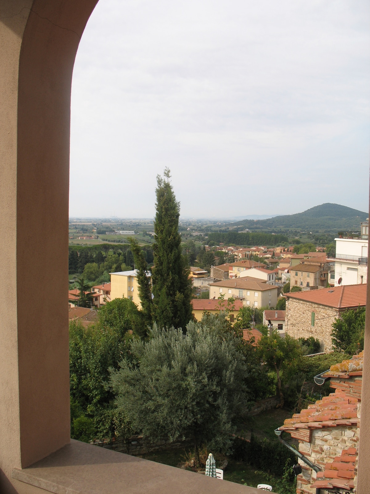 A view from an arched window showcases a lush valley with rolling hills in the distance. Green trees and residential buildings are visible below, along with a tiled roof in the foreground. The overall scene offers a glimpse of the surrounding natural landscape.
