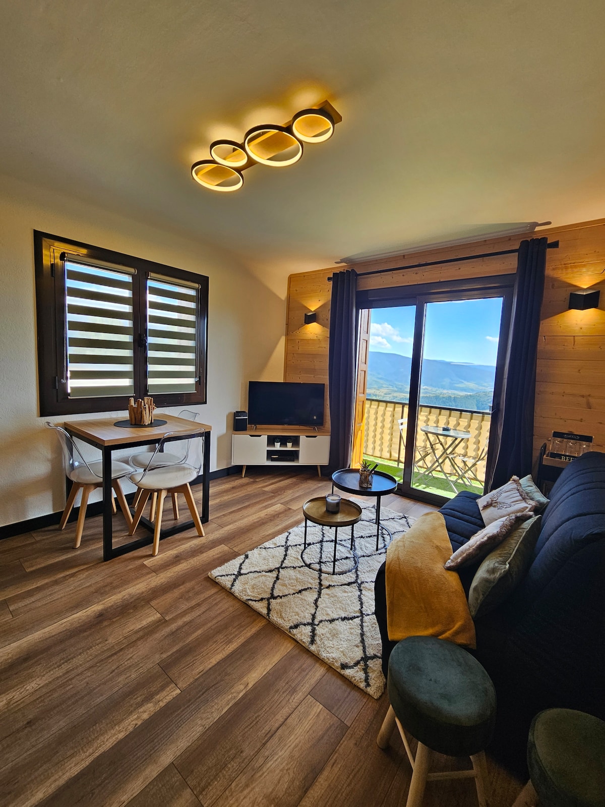 A bright living area features a dark sofa and a small dining table with light wooden chairs. A television is positioned against the wall, and large glass doors open to a balcony with mountain views. Natural light enhances the warm wood textures.