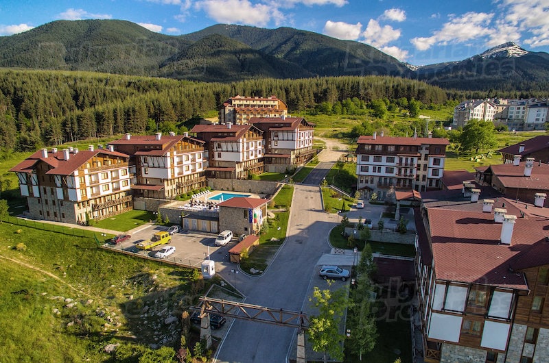 Aerial view of a modern complex surrounded by evergreen trees and mountains. The layout includes several buildings with a central outdoor pool visible, along with a road leading through the development. Scenic views of nature extend in all directions.