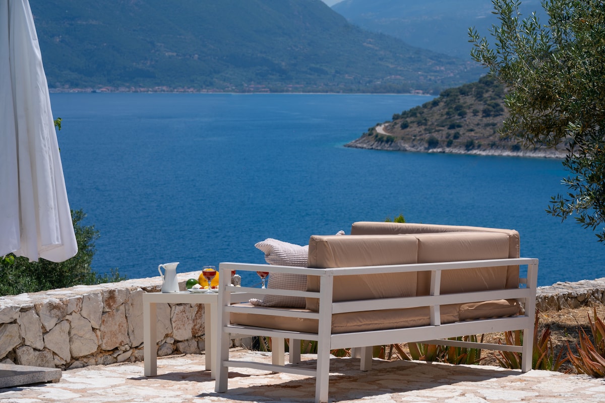 A spacious outdoor seating area is set against a stunning backdrop of the sea and mountains. A light-colored sofa with neutral cushions faces the water, inviting relaxation. A small table nearby holds refreshments, complementing the serene coastal views.