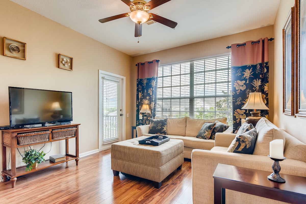 A comfortable living area features a large sectional sofa arranged around a coffee table. A flat-screen TV is positioned on a wooden console. Natural light fills the space through multiple windows, which are dressed with elegant curtains, adding warmth to the room.