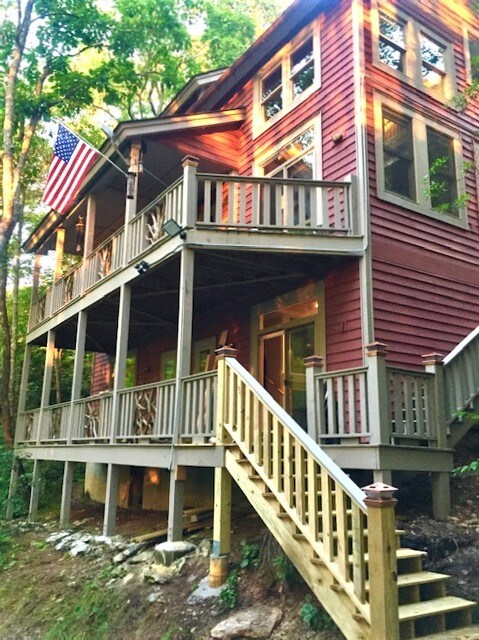 The exterior of a three-story craftsman-style mountain home is showcased, featuring a wraparound porch and wooden railings. An American flag is proudly displayed, while sunlight filters through surrounding trees, highlighting the warm red tones of the house.