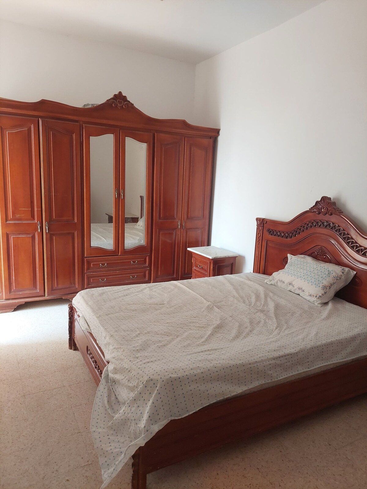 A serene bedroom is furnished with a wooden bed and a matching wardrobe featuring mirrored doors. Natural light enters the room, highlighting the textured bedding. A small bedside table stands next to the bed, contributing to the room's calm and inviting ambiance.