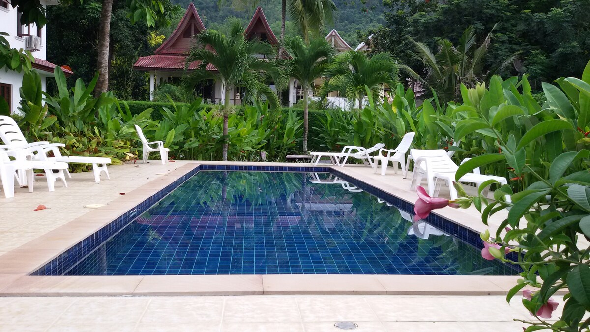 A community pool surrounded by lush greenery and tropical plants is depicted. White lounge chairs are positioned at the poolside. The tranquil water reflects the surrounding flora and the architecture of nearby structures in the background.