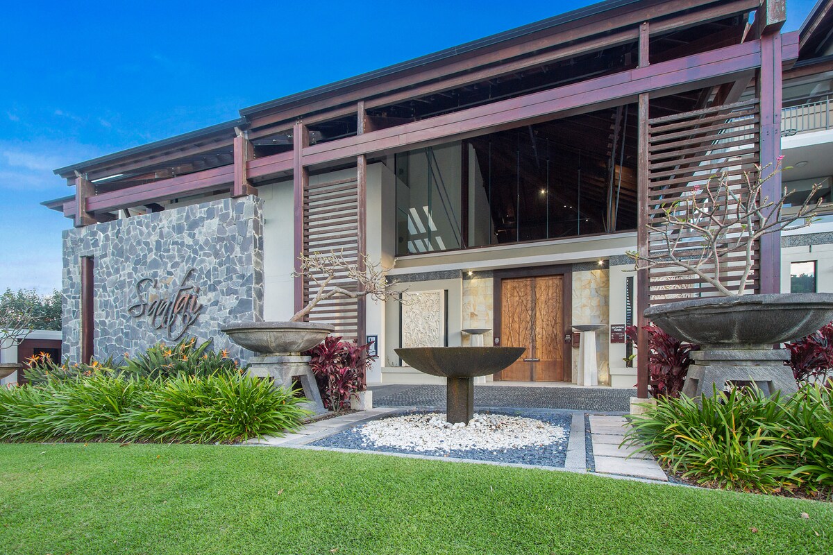 The entrance features a modern and striking facade, accented by natural stone and warm wood elements. Smooth stone walkways lead to a grand entrance, flanked by decorative planters and lush greenery, creating an inviting atmosphere for guests.