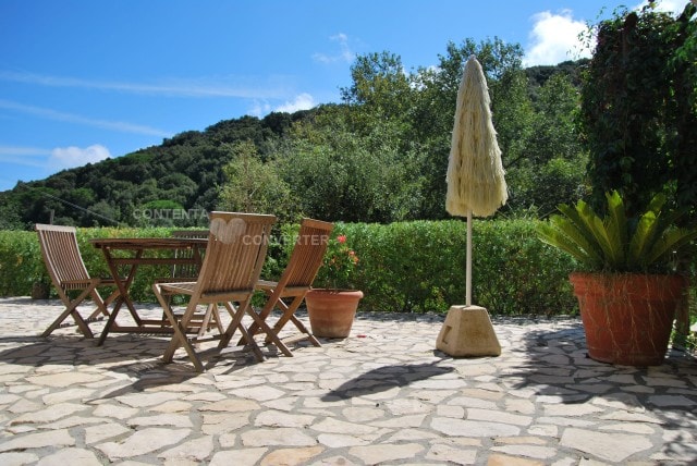 An outdoor terrace features a wooden dining table surrounded by four folding chairs, complemented by a freestanding umbrella for shade. Lush greenery and a low stone wall are visible in the background, creating a natural setting with a clear blue sky above.