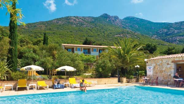 A large swimming pool is surrounded by palm trees and comfortable lounge chairs. Umbrellas provide shade for guests enjoying the outdoor space. A mountainous landscape rises in the background, complementing the serene ambiance of the area.
