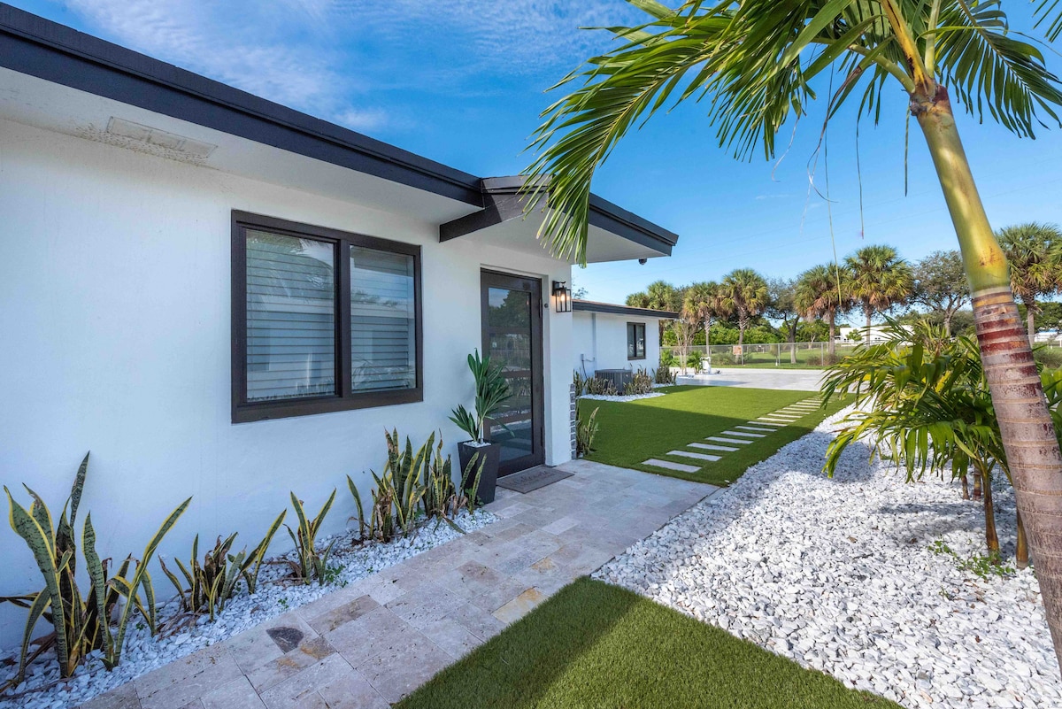 The entrance features a modern facade with a large glass door and clean lines. Lush green landscaping is complemented by neatly arranged white stones along the pathway, leading to a vibrant patch of grass and palm trees in the background.
