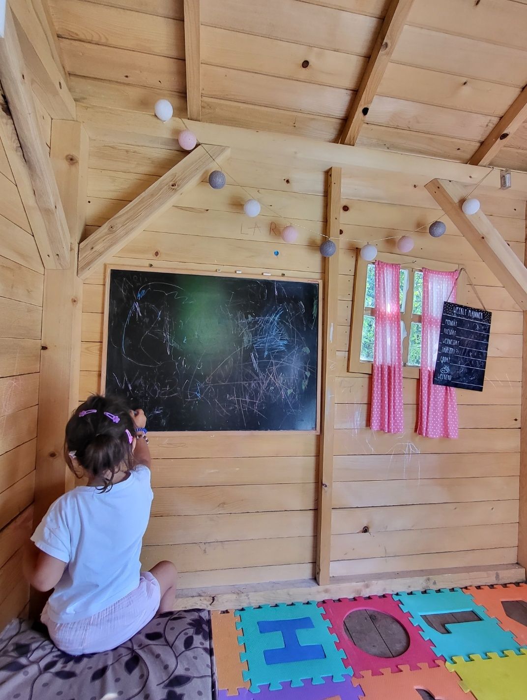 A charming playroom is showcased, featuring a black chalkboard on a wooden wall, where a child is drawing. Colorful foam floor tiles provide a soft surface. Curtains frame a window, and decorative garlands add a playful touch to the space.