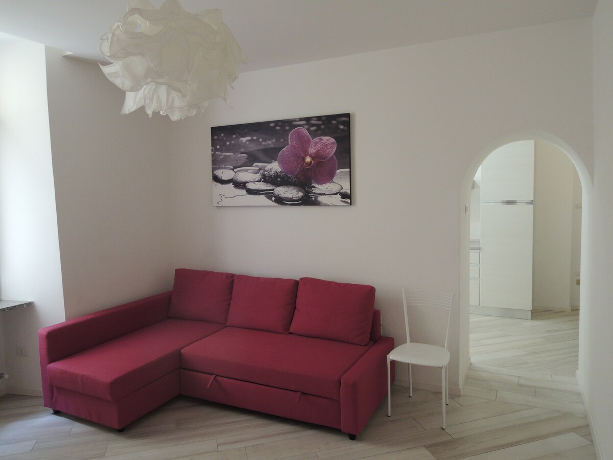 A corner seating area is furnished with a red sectional sofa, providing a cozy spot for relaxation. A white chair is positioned nearby, while a floral artwork featuring an orchid adds a touch of elegance to the wall. An archway leads to the kitchen area.