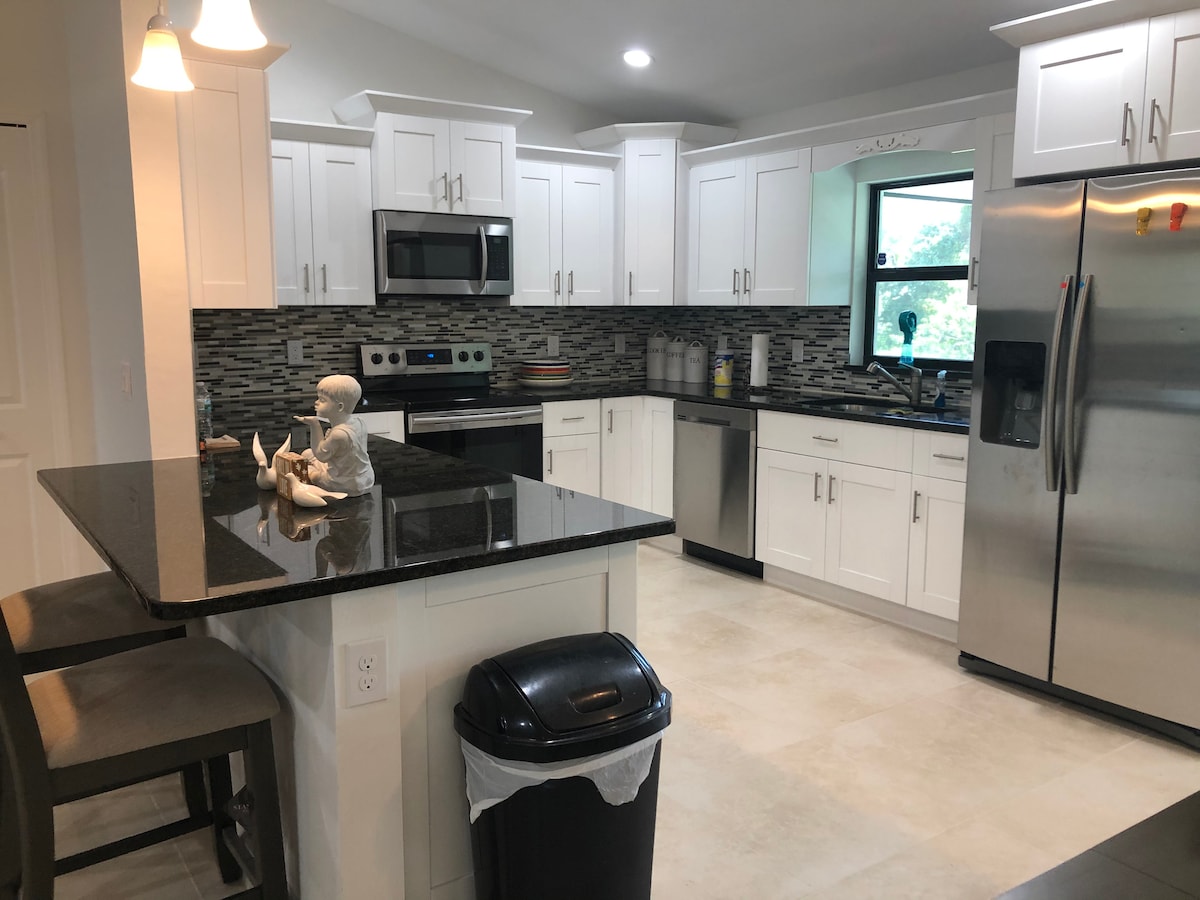 A modern kitchen is equipped with stainless steel appliances and white cabinetry. A dark granite countertop extends into a small eating area with two bar stools. Light-colored tiles cover the floor, and a window offers a view to greenery outside.