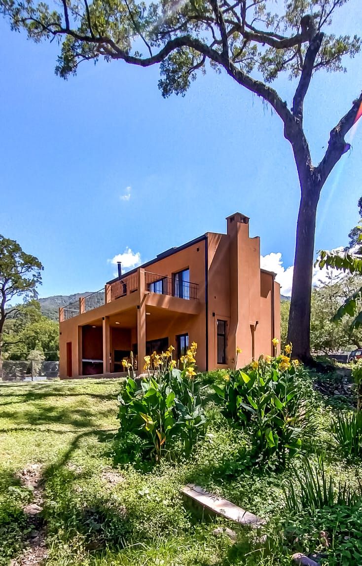 A modern two-story house is set against a backdrop of lush greenery. Large windows provide natural light, while a wide balcony extends from the upper floor. Vibrant yellow flowers and well-maintained grass surround the exterior, contributing to the peaceful outdoor setting.