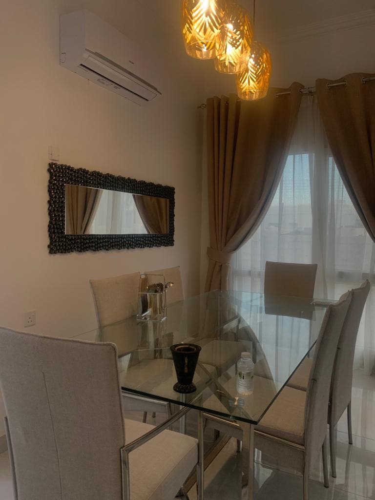 A modern dining area features a sleek glass table accompanied by six soft, upholstered chairs. Elegant lighting fixtures hang overhead, illuminating the space. A large mirror is mounted on the wall, reflecting the soft light from the windows dressed with beige curtains.