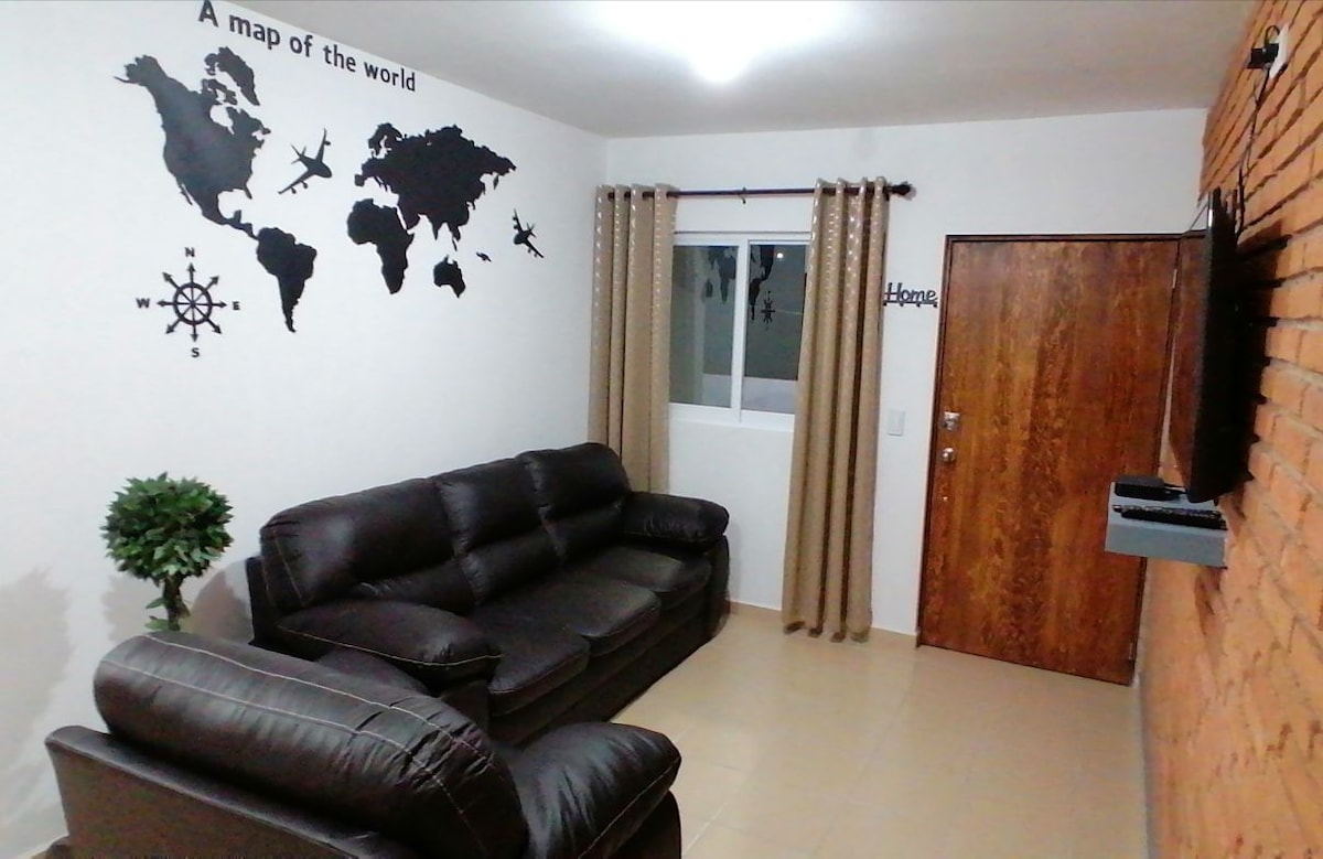 A cozy living area features a dark leather sofa set, complemented by decorative wall art displaying a world map. Natural light enters through a nearby window, surrounded by light-colored curtains, enhancing the space's welcoming feel. A flat-screen television is mounted on a textured wall.