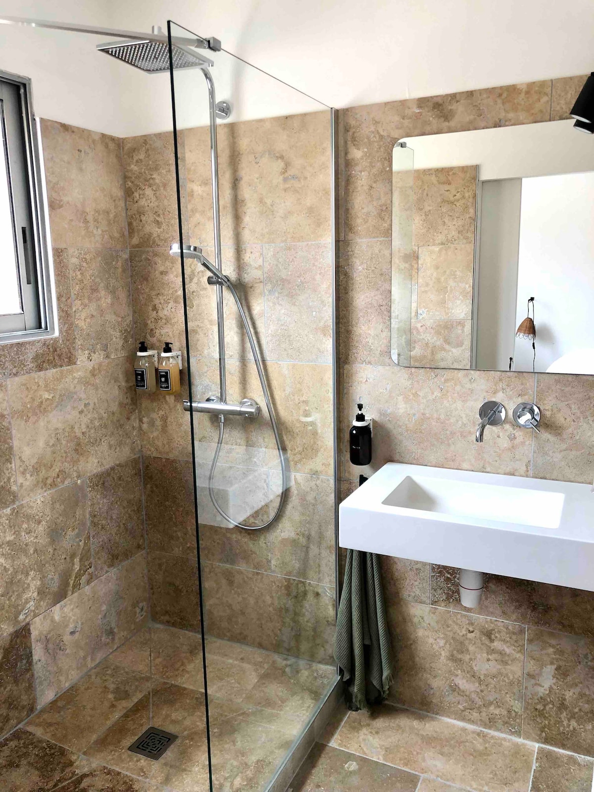 The spacious bathroom features a walk-in shower enclosed by clear glass. Natural stone tiles in earthy tones cover the walls and floor. A modern sink with a minimalist design is placed beneath a mirror, while toiletries are displayed on the adjacent wall.