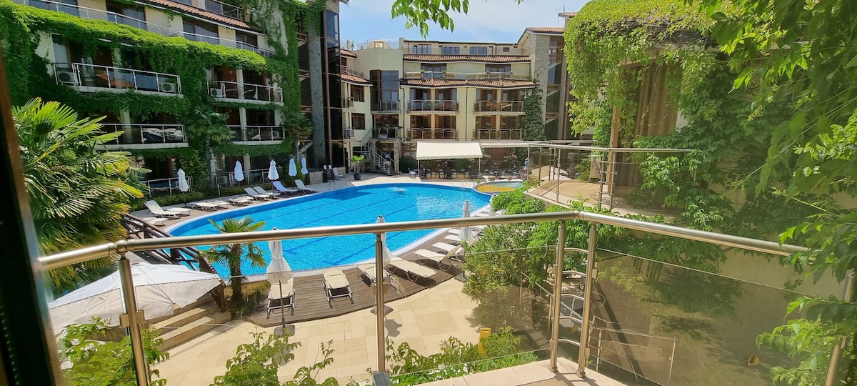 A view of the swimming pool area is presented from a spacious terrace. The pool is surrounded by lounge chairs and palm trees, with the buildings of the resort visible in the background, adorned with greenery. Sunlight illuminates the scene, creating a serene atmosphere.