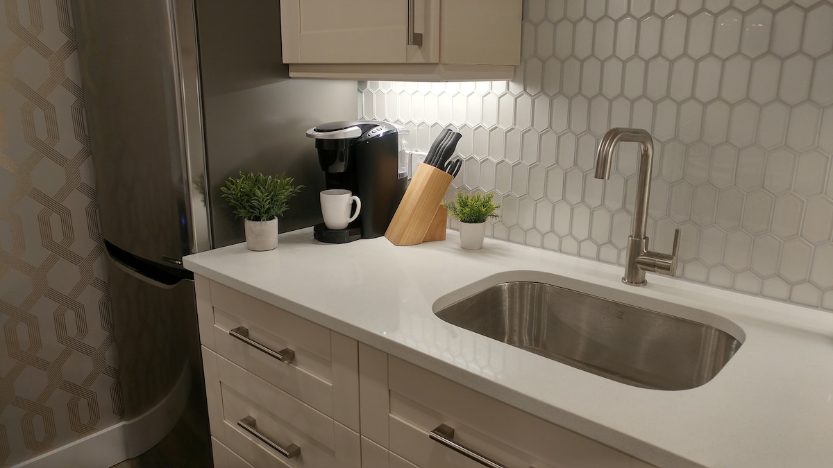 A modern kitchen area features a white countertop and a stainless steel sink. Hexagonal white tiles create a stylish backsplash. A Keurig coffee machine sits atop the counter, accompanied by cutlery in a wooden holder and small potted plants for a touch of greenery.