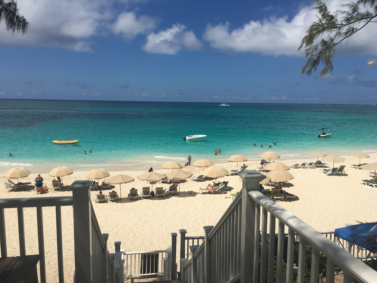 A view of a sandy beach is presented, where sun umbrellas and lounge chairs are spaced along the shore. Calm turquoise waters stretch into the distance, with a few boats seen on the horizon. A clear blue sky is dotted with clouds, contributing to a serene coastal setting.