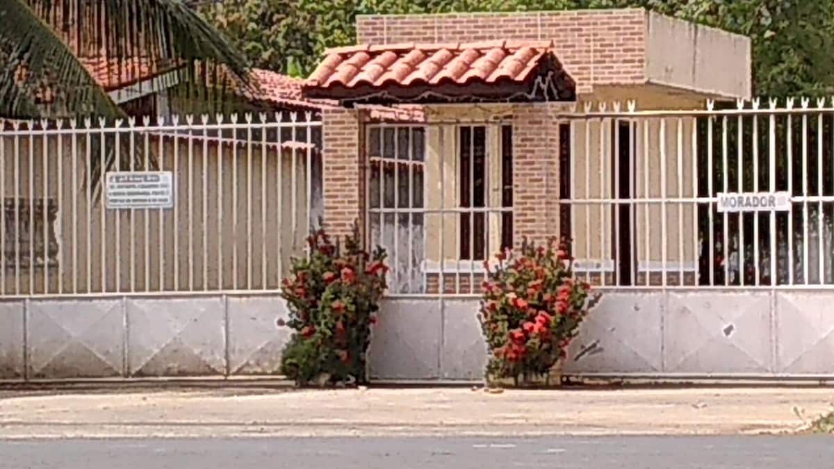 The entrance to the property is framed by a decorative gate with a brick archway. Vibrant red flowering plants are positioned symmetrically on either side, enhancing the welcoming appearance of the entrance. The building beyond is partially visible through the gate.
