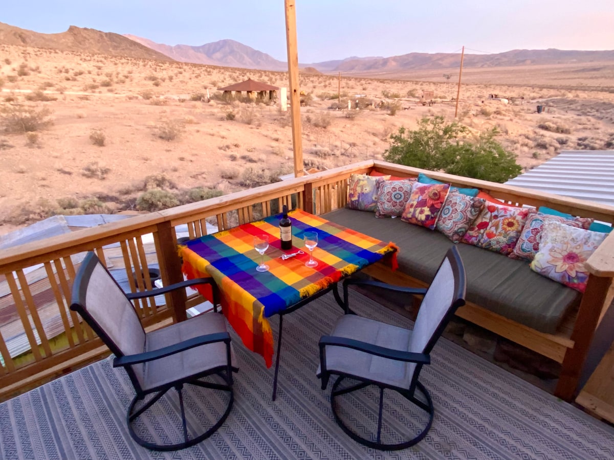 An outdoor deck features a colorful table with two glasses, complemented by a vibrant tablecloth. A comfortable seating area is adorned with cushions. The surrounding desert landscape and mountains provide an expansive backdrop, contributing to a serene atmosphere.
