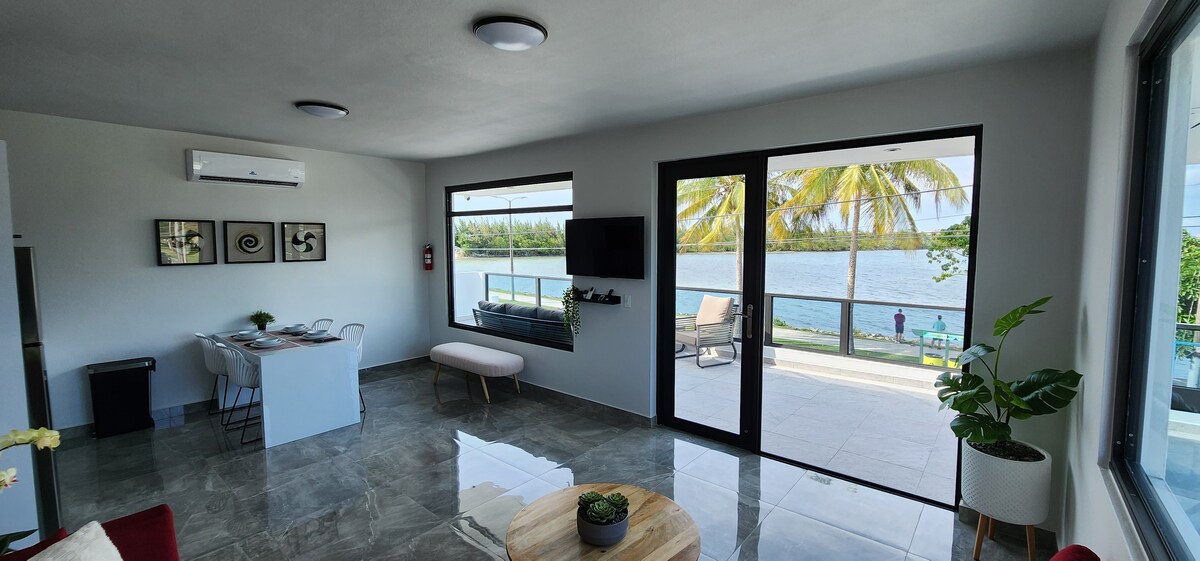 A spacious living area features large windows that offer views of the bay. A small dining table is set nearby, while a light-colored bench sits in front of the glass doors. Outdoor lounging chairs are visible on the patio, surrounded by palm trees.