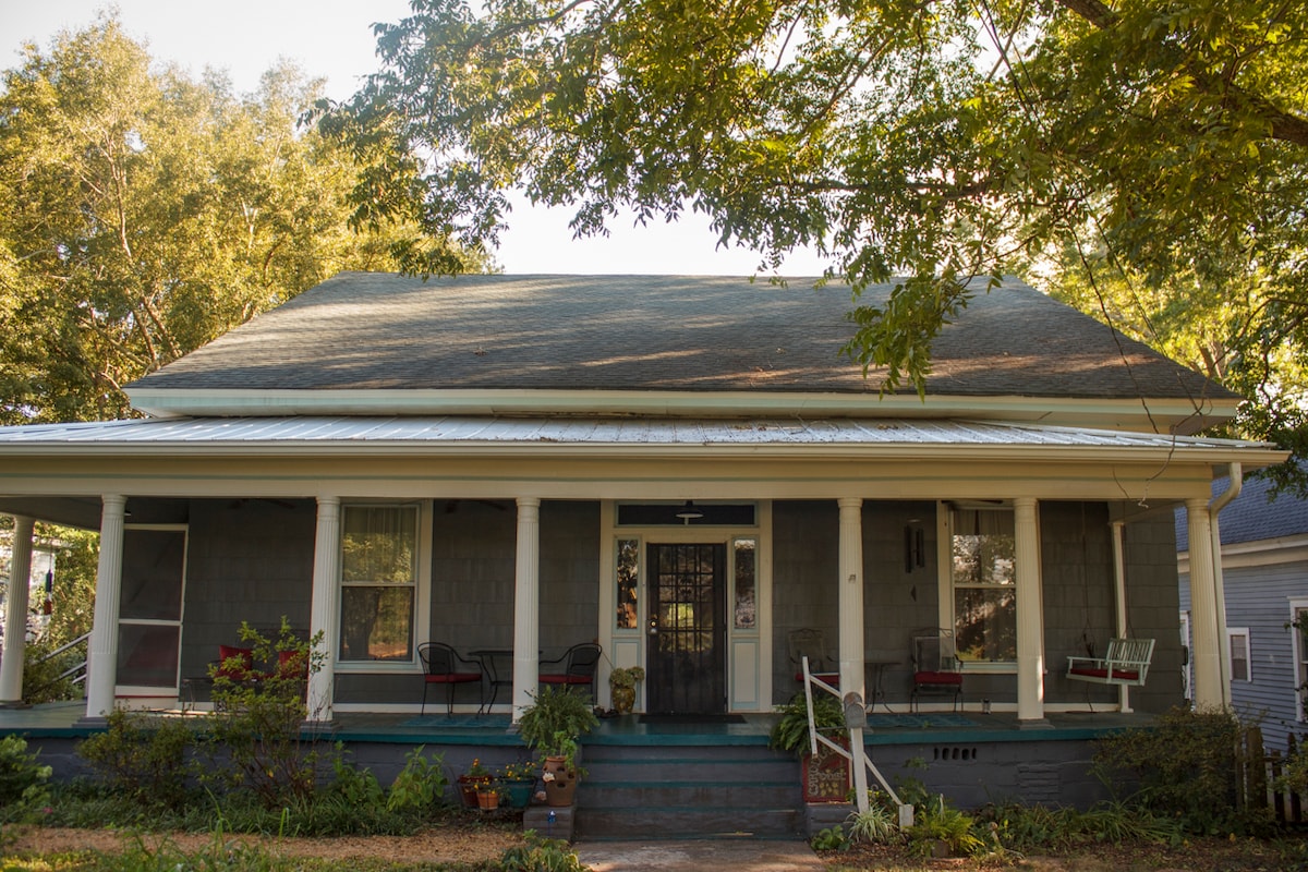 The exterior of a two-story gray house is shown, featuring a spacious porch with white columns. Comfortable seating is arranged on the porch, surrounded by greenery and flower pots. Large trees provide shade over the front yard, creating a welcoming appearance.