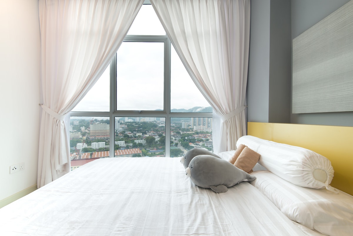 A bright bedroom area features a spacious bed with soft linens and decorative pillows. Large windows allow natural light to fill the room, showcasing views of the cityscape and distant hills. Light-colored curtains frame the windows, creating a calming atmosphere.