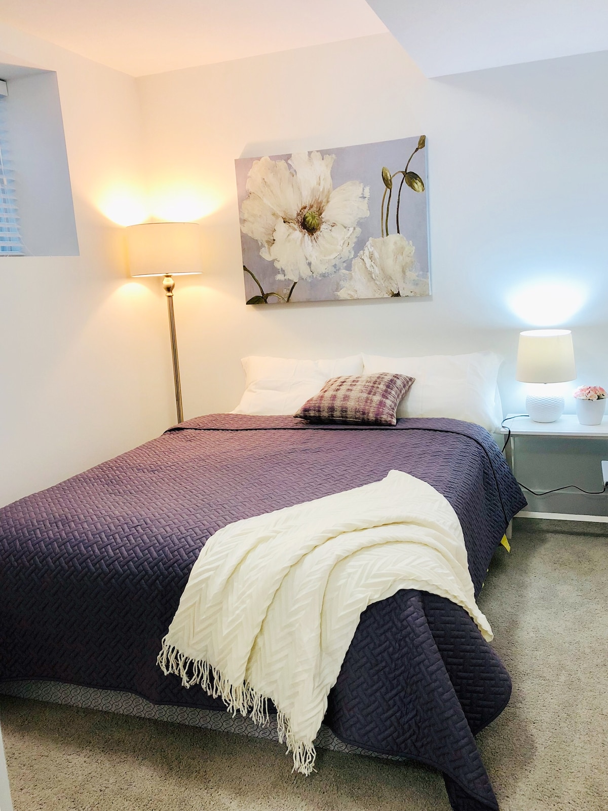 A cozy bedroom features a queen size bed adorned with a dark purple blanket and a patterned throw pillow. Soft lighting is provided by two bedside lamps, while a floral artwork is displayed on the wall above the bed. A warm, inviting atmosphere is created with neutral tones.