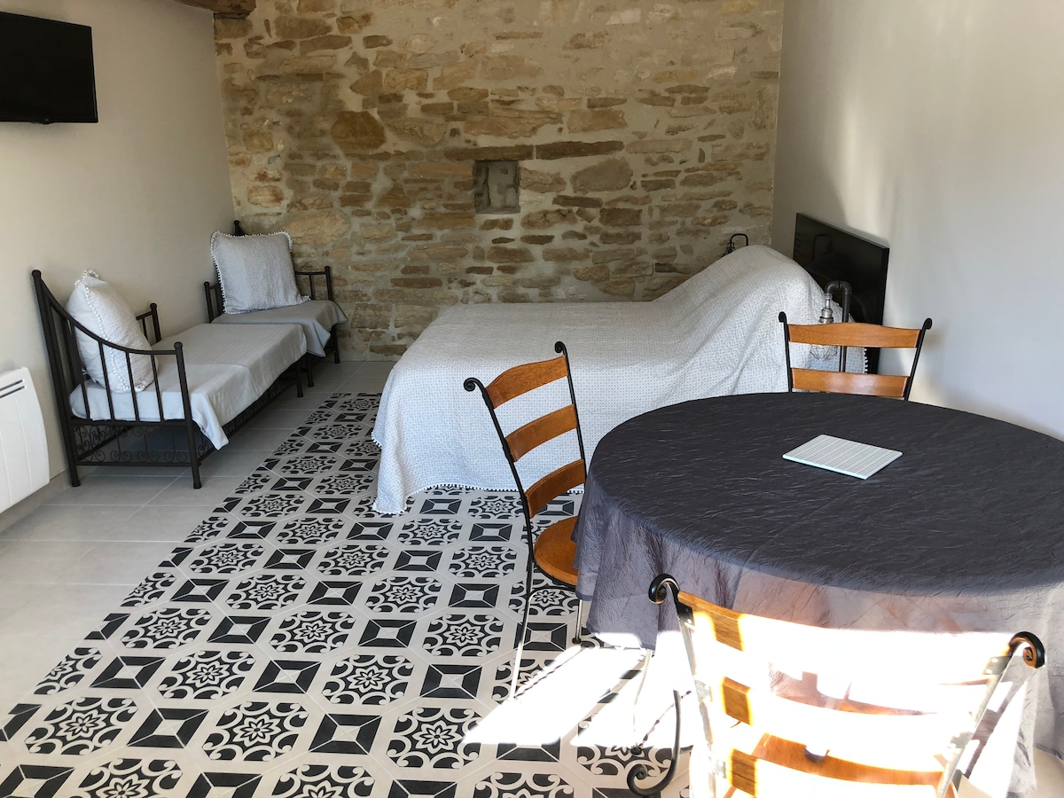 A spacious room features a textured stone wall and a cozy double bed covered with a light quilt. A round table with a dark cloth is accompanied by wooden chairs, while additional seating is provided by two lounge chairs in the corner. Natural light fills the space.