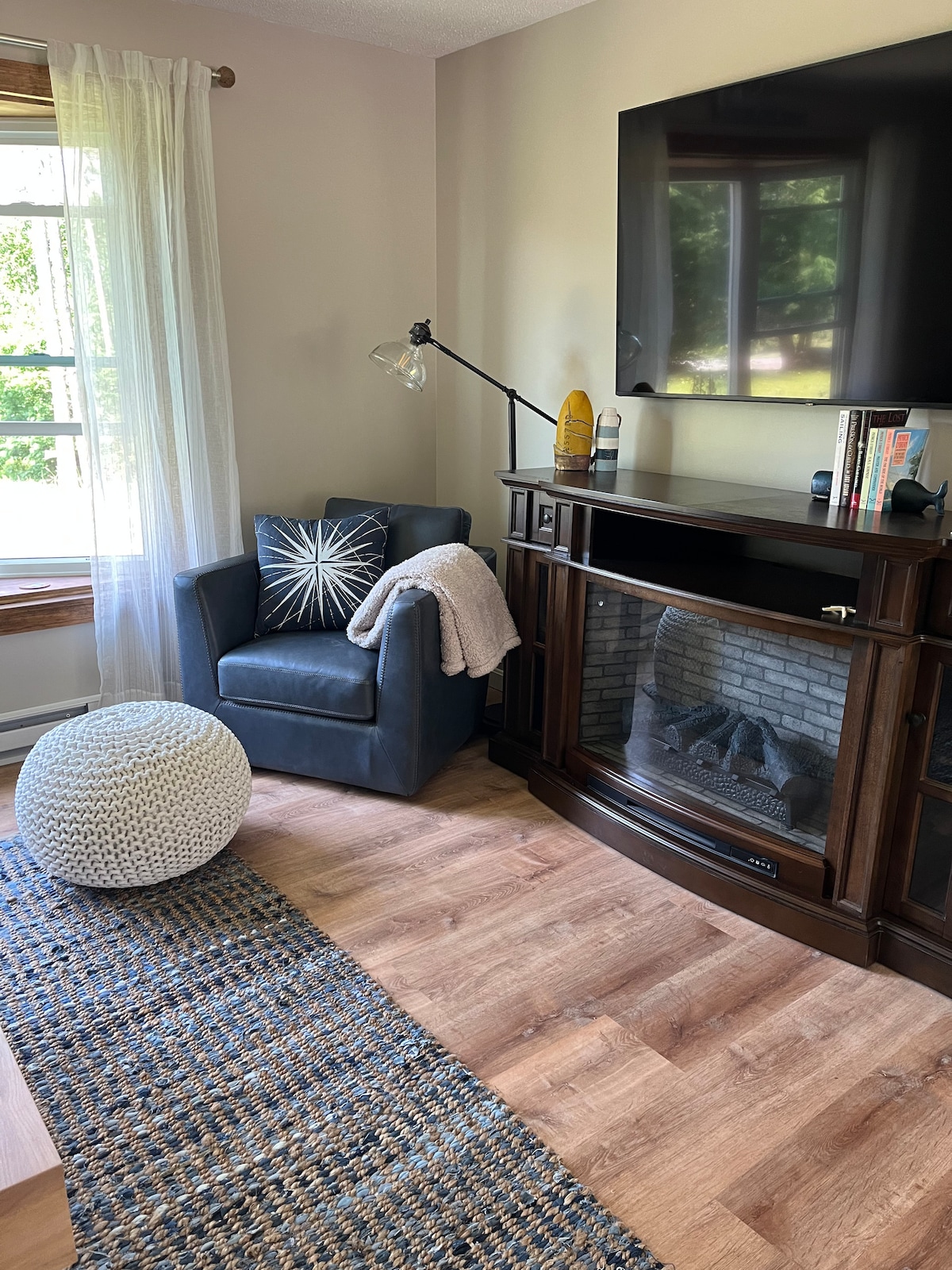 A cozy seating area features a blue armchair adorned with a decorative pillow and a soft throw. A small pouf is positioned nearby, while a television is mounted above a wooden console with a glass-front fireplace. Natural light fills the space through large windows with sheer curtains.