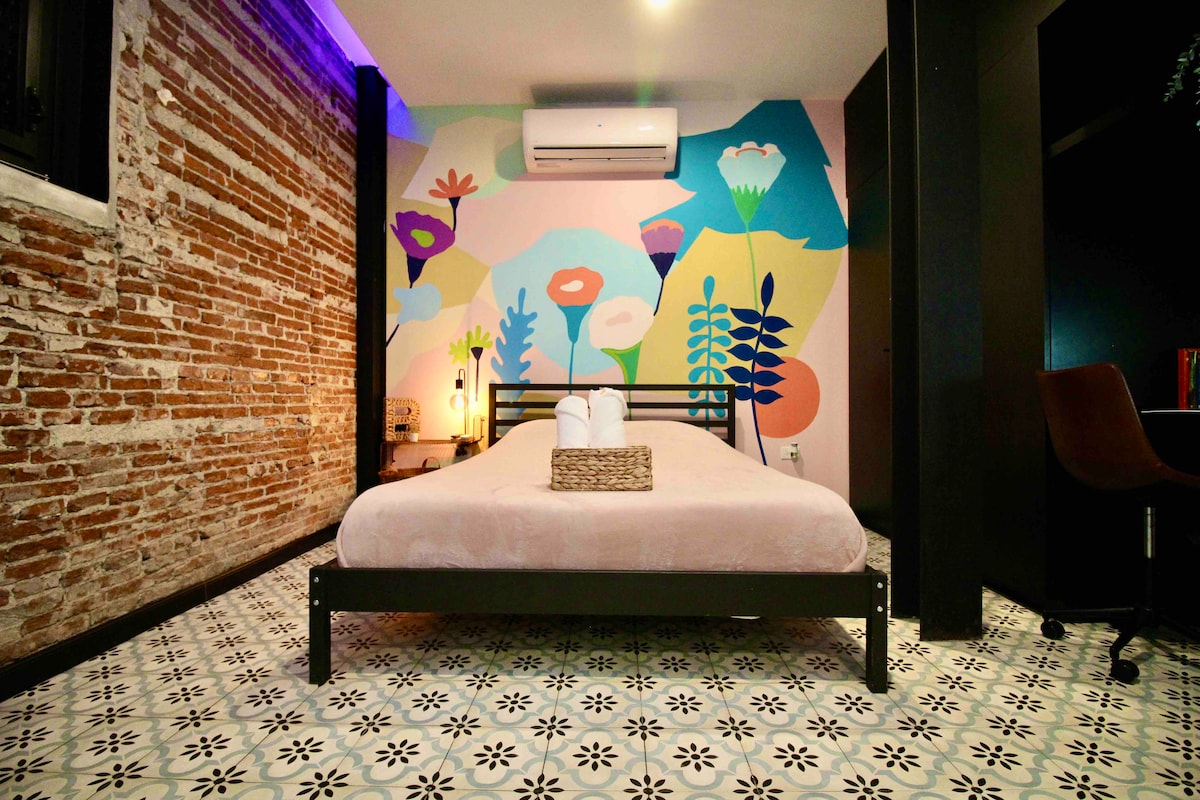 A studio-style apartment features a queen-size bed with a light pink coverlet, positioned against a vibrant mural depicting colorful flowers and foliage. Exposed brick walls add texture, while decorative tiles cover the floor. Soft lighting creates a cozy ambiance.