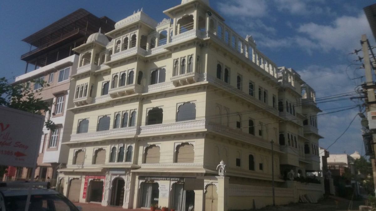 The heritage-style building features intricate architectural details with multiple arched windows and balconies. Its elegant facade is adorned with traditional motifs, showcasing charming elements characteristic of local design. The structure stands against a bright blue sky, enhancing its prominent presence in the area.