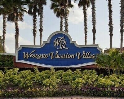 A welcoming sign for Westgate Vacation Villas is prominently displayed, surrounded by lush greenery and palm trees. The sign features the initials 'WR' and is set against a blue background, conveying a sense of relaxation and vacation spirit.