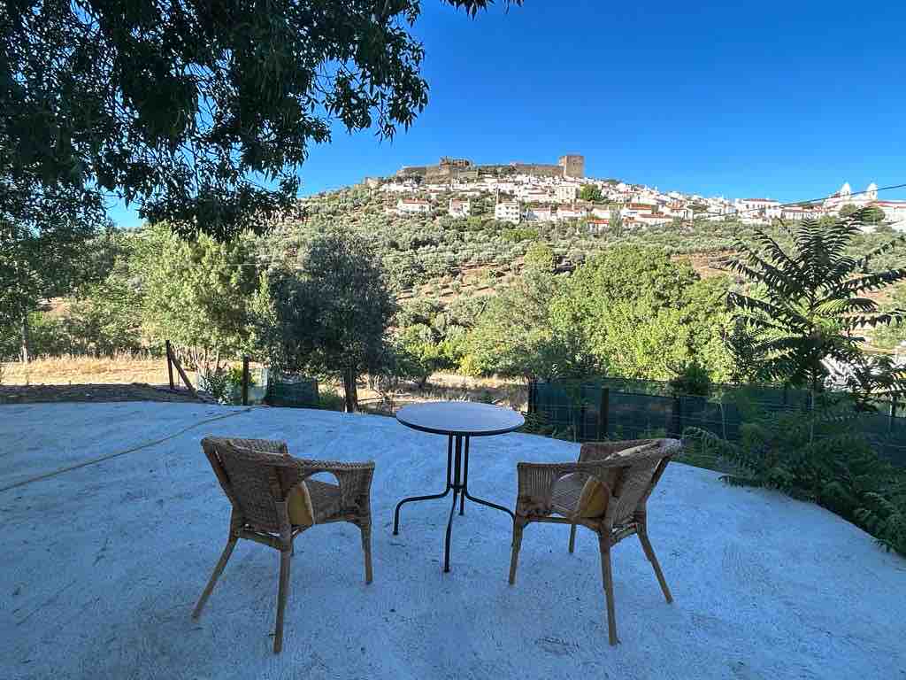 A small outdoor seating area is featured, consisting of two woven chairs positioned around a circular table. The background showcases a hillside village and castle, surrounded by lush greenery and trees under a clear blue sky.