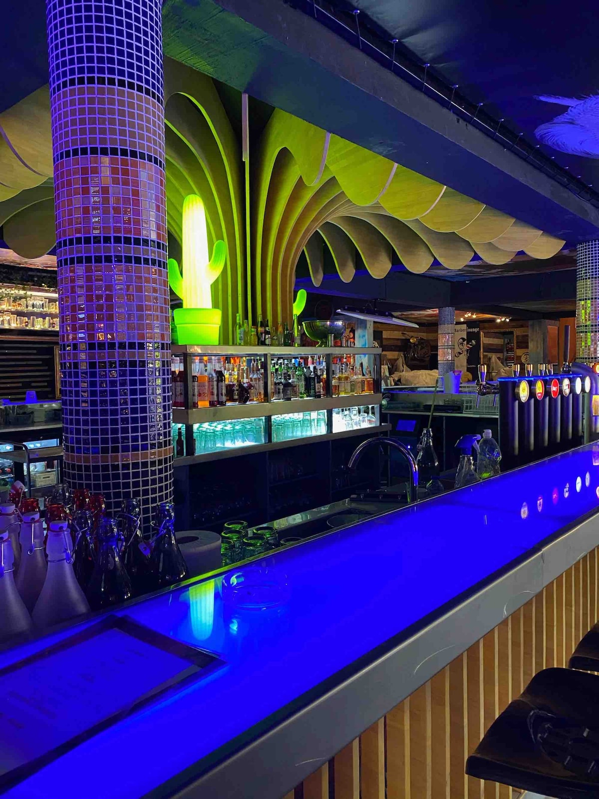 A vibrant bar area is highlighted by colorful lighting. The counter features a sleek, illuminated surface in blue. Shelves filled with various bottles are visible behind the counter, while unique decorative elements, such as a neon-green cactus and large foliage-inspired designs, enhance the lively atmosphere.